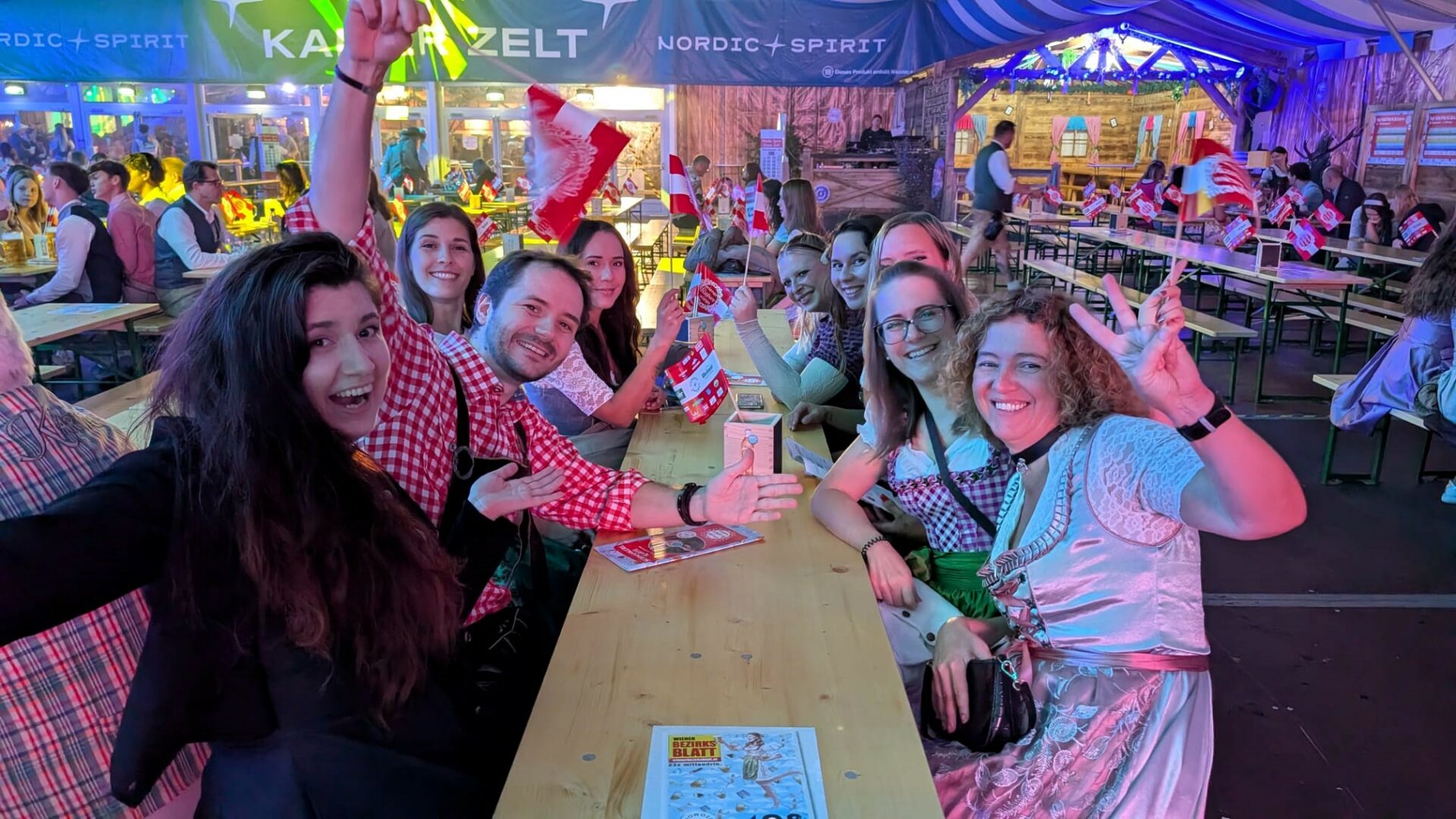 Eine Gruppe von Menschen sitzt auf Heurigenbänken in einem Festzelt, einige schwingen Fahnen.